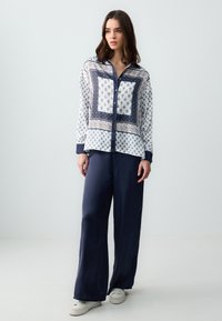 Chemise à boutons ornée d'un motif paisley bleu marine et blanc avec une bordure accentuée, associée à un pantalon satiné bleu marine ample et des baskets blanches.