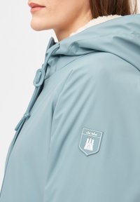 Chaqueta impermeable azul con una tela texturizada, que cuenta con una capucha forrada de piel sintética, cordón ajustable y un logo de parche en la manga.