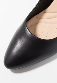 Chaussure plate en cuir noir à bout pointu avec semelle intérieure perforée et rembourrée beige, conçue pour le confort.