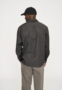 Schwarz- und grau gestreiftes Langarmhemd mit Knopfmanschetten, ausgestattet mit einem klassischen Kragen und einem Rückeneinsatzdesign. Kombiniert mit hellgrauen Hosen.