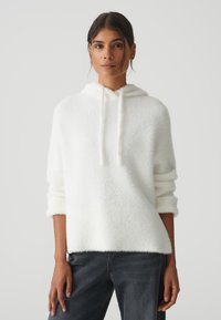 Weißer Kapuzenpullover aus weichem, strukturiertem Material. Er hat eine lockere Passform und aufgerollte Ärmel und wird mit dunkelgrauen Jeans kombiniert.