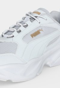 Λευκό και γκρι αθλητικό παπούτσι Puma με χρυσό λογότυπο στο πλάι και στη γλώσσα, άνω μέρος από δίχτυ και δέρμα και χοντρή λευκή σόλα.