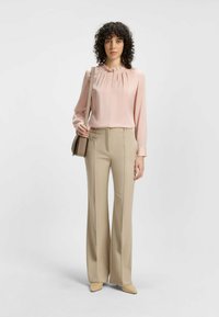 Blusa a maniche lunghe rosa chiaro con scollatura plissettata, abbinata a pantaloni beige a campana. Il modello tiene una piccola borsa a tracolla beige.