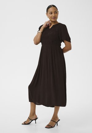 Kobieta stoi z ręką przy twarzy, ubrana w czarną sukienkę do połowy łydki i czarne sandały na obcasie na jednolitym białym tle.