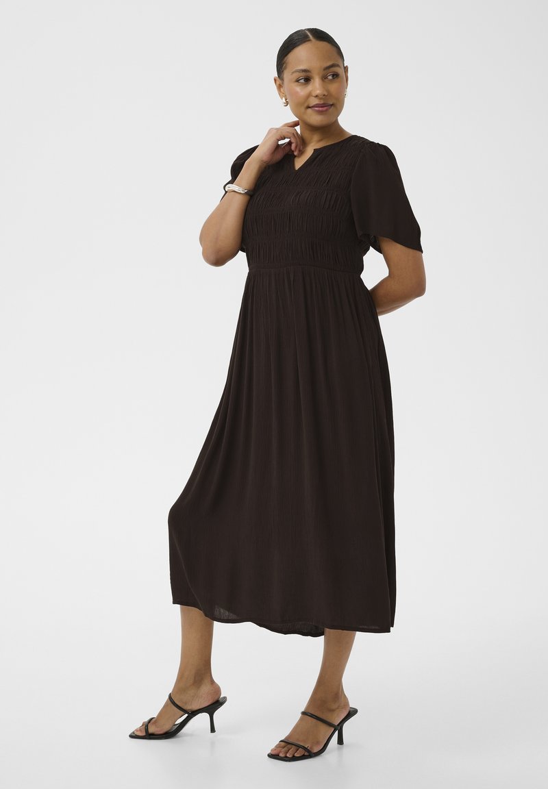 Femme debout, main près du visage, portant une robe noire longueur mollet et des sandales à talons noires sur un fond blanc uni.