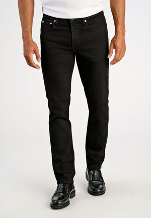 Schwarze Jeans aus Denim mit schlanker Passform, ausgestattet mit kupferfarbenen Nieten und einem klassischen Fünf-Taschen-Design, kombiniert mit schwarzen Slippern.