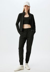 Zwarte sportoutfit bestaande uit een aansluitende zip-up jack en hoogsluitende broek, gemaakt van een gladde, rekbare stof. Inclusief een lichte pet en witte sneakers.