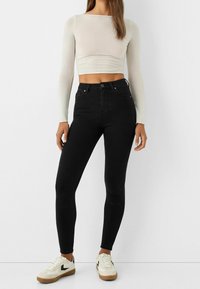 Mujer vestida con jeans ajustados de cintura alta negros, top corto de manga larga beige y zapatillas blancas con detalles en negro y marrón, de pie.