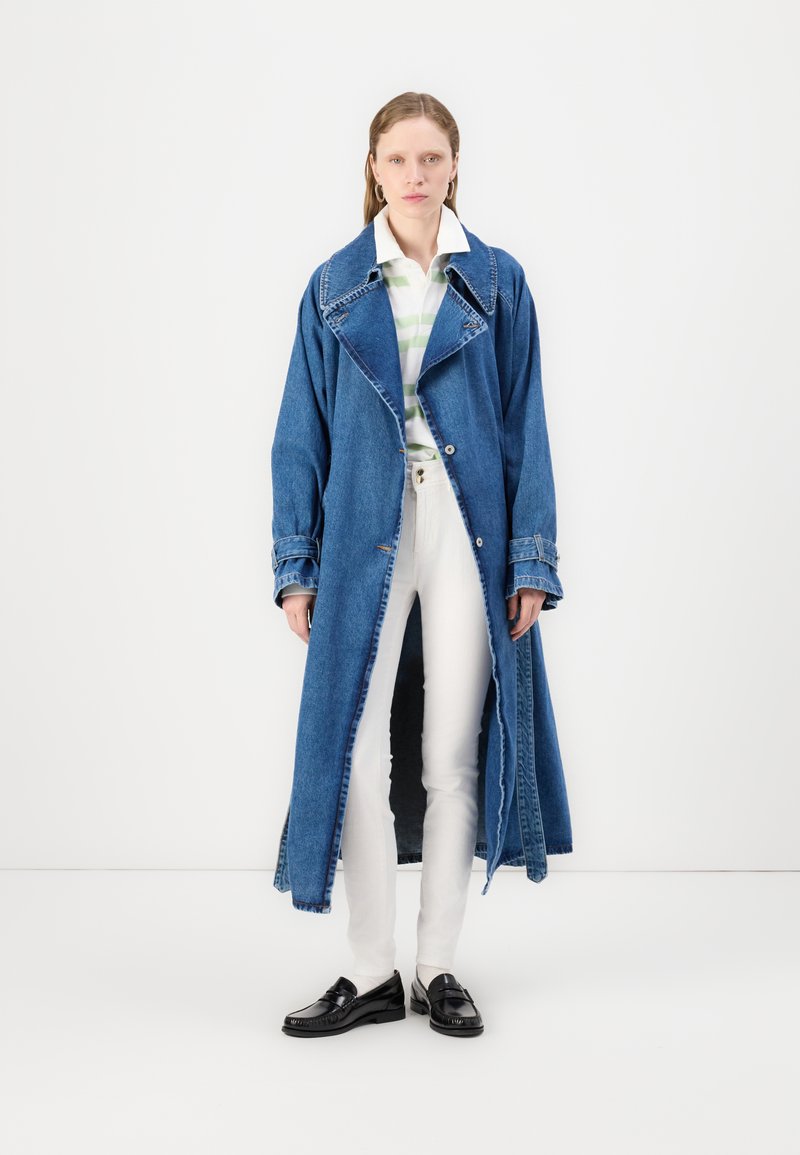 Denim-Jacke mit langem, lockeren Schnitt, weitem Kragen und Kontrastnähten. Getragen über einem gestreiften Hemd und weißen Hosen, kombiniert mit schwarzen Schuhen.