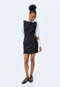Schwarzes, tailliertes Kleid mit ärmellosem Design, über einem weißen Button-Down-Hemd getragen. Glattes Material, knielang, kombiniert mit schwarzen, glänzenden Schuhen.