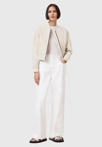 Chaqueta bomber de color beige con cremallera frontal, llevada sobre una camiseta gris clara, combinada con pantalones de pierna ancha blancos de cintura alta y sandalias negras.