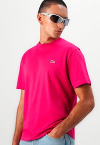 Fuchsia bomulds t-shirt med rund halsudskæring, korte ærmer og lille grøn logo på brystet. Båret med grå solbriller.
