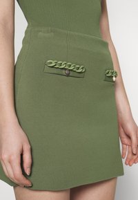 Mini-jupe verte en tricot avec deux poches avant ornées de détails tressés et de boutons métalliques. Texture lisse avec une coupe ajustée.