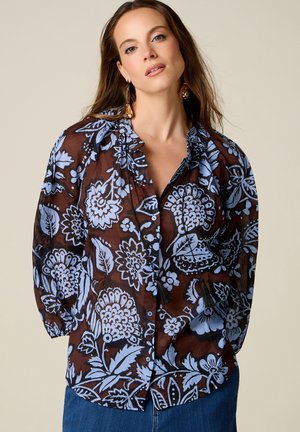 Vrouw met lang bruin haar die gouden oorbellen draagt en een bruine blouse met grote lichtblauwe bloemenprints en een blauwe spijkerbroek.