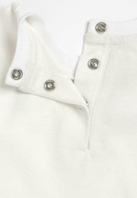 Vêtement en coton blanc avec une patte de boutonnage à pression au niveau de l'encolure, muni de pressions en métal argenté et d'une texture lisse.