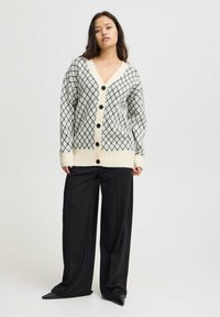 Cardigan a bottoni color crema con motivo a rombi neri, con maniche lunghe e orlo a coste. Indossato con pantaloni neri tailleur e scarpe con punta.