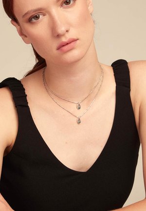 Colliers en argent superposés avec des pendentifs ovales. Le mannequin porte un haut sans manches noir avec des bretelles volantées. L'arrière-plan neutre met en valeur le sujet.