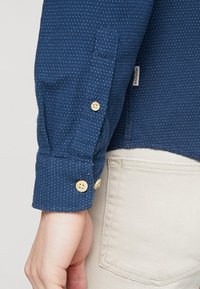 Tummansininen paita, jossa pieniä valkoisia pilkkuja, pitkät hihat, napilliset kalvosimet ja hillitty tekstuurikangas. Päällä vaaleiden housujen kanssa.