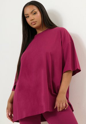 Mujer joven con cabello largo y oscuro, vestida con camiseta magenta de gran tamaño y leggings a juego, de pie frente a una pared blanca.