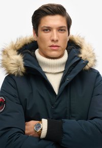 Mørkeblå dunjakke med en pelsforet hætte, der har et logo patch på ærmet og en ribbet krave. Bæres over en cremefarvet turtleneck.