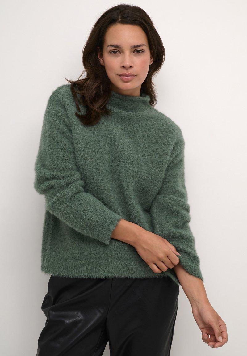 Grüner, flauschiger Pullover mit hohem Hals, lockerer Passform und langen Ärmeln. Weiche Textur und leicht gerippter Saum, kombiniert mit schwarzen Lederhosen.
