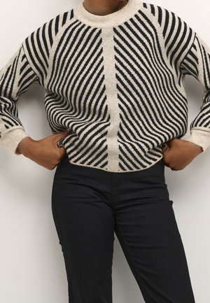 Osoba nosící béžovo-černý pruhovaný svetr s geometrickým vzorem, v kombinaci s černými kalhotami, s rukama opřenýma v bok.