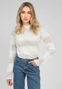 Un haut en dentelle blanche avec un design à motifs et des découpes rayées, des manches longues et un col montant, assorti à un jean en denim bleu taille haute.