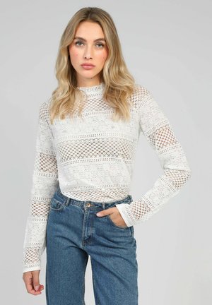 Un haut en dentelle blanche avec un design à motifs et des découpes rayées, des manches longues et un col montant, assorti à un jean en denim bleu taille haute.