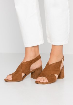 Brown suede open-toe heels with a block heel, featuring crossed straps and a slingback design with adjustable buckle. Neutral-colored sole.