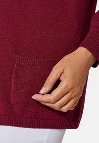 Pull en tricot bordeaux avec un col et des poignets côtelés, orné d'une petite poche avant visible. Texture lisse et coupe décontractée.