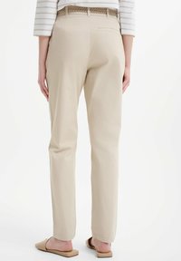 Beżowe spodnie o gładkiej fakturze, prostej linii i plecionym pasku. Posiadają tylnią kieszeń i są noszone z płaskimi, jasnymi sandałami.