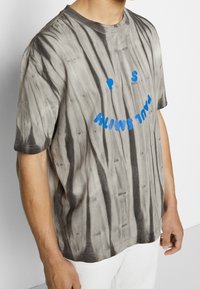 Camiseta de manga corta gris con estampado tie-dye y acabado texturizado. Presenta texto impreso en azul, incluyendo una carita sonriente y la frase "PAUL SMITH."