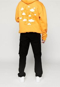 Oranžová mikina s kapucí s bílým motivem mraku na zádech, doplněná černými cargo kalhotami a bílými teniskami, nabízející volný střih.