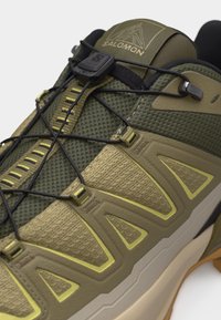 Κοντινή λήψη από παπούτσι για τρέξιμο σε μονοπάτια σε ελιά πράσινο χρώμα με μαύρο σύστημα γρήγορου δέσιμου, υφασμάτινο επάνω μέρος με υφή και μπεζ λαστιχένια σόλα.