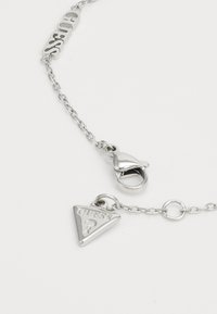 Silver chain necklace featuring a triangular charm with the text "GUESS?" and a clasp, showcasing a sleek metal design.