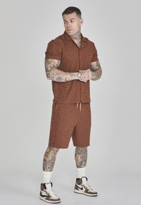 Braunes Kurzarmhemd und passende Shorts mit geprägtem Muster; getragen mit weißen Socken und braunen High-Top-Sneakern.