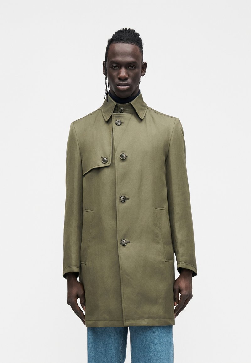 Olive green trench coat with a collar, four button closure, and two side pockets. Smooth texture with a tailored fit, worn over a black turtleneck.
