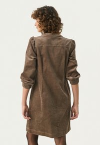 Ruskea corduroy-mekko, jossa on ilmapallohihoja, pyöreä kaulus ja polven pituinen helma. Kankaassa on ribbipinta ja rento istuvuus.