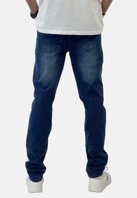 Jeans de denim azul oscuro con un corte ajustado, que presentan bolsillos traseros y un ligero desgaste. Combinados con zapatillas blancas, vistos desde la parte de atrás.