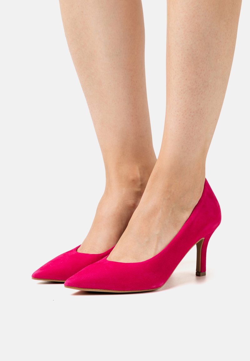 Femme portant des escarpins en daim rose vif à bout pointu avec talons moyens et fins sur fond blanc.