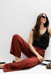 Débardeur noir côtelé, pantalon rouge texturé ample et sandales noires ; tenant un sac noir et beige. Pose assise décontractée contre un mur blanc.