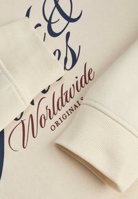 Tela de color crema con puños acanalados, que presenta texto impreso en azul marino y burdeos. El diseño incluye "Worldwide Originals" en una tipografía elegante.