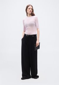 Γυναίκα με μακριά καστανά μαλλιά φοράει ροζ ανοιχτό ριπτό κοντομάνικο τοπ, φαρδιά μαύρη παντελόνια με ζώνη, μαύρα παπούτσια και κρατάει ένα μαύρο τσαντάκι.