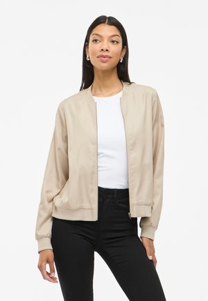 Beige bomber jacket featuring a smooth texture, front zip closure, ribbed cuffs and hem, worn over a white top and black jeans.