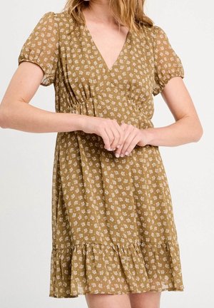 Femme portant une robe jaune moutarde à manches courtes avec de petites impressions florales blanches et un ourlet volanté, debout les mains jointes.