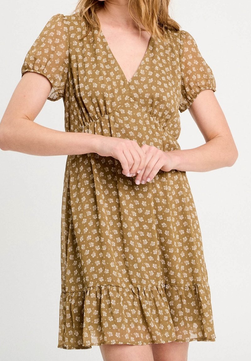 Femme portant une robe jaune moutarde à manches courtes avec de petites impressions florales blanches et un ourlet volanté, debout les mains jointes.