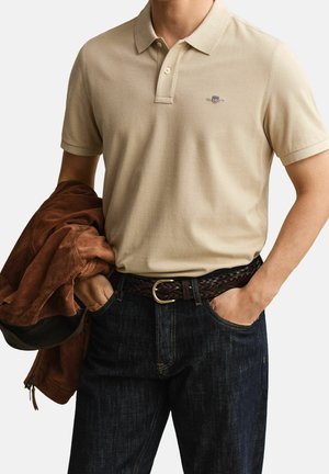 Mand iført beige kortærmet polo shirt stoppet ind i mørke jeans med flettet brunt bælte, der holder en brun jakke i den ene hånd.