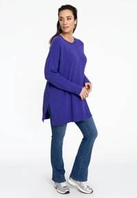 Lila Langarmpullover mit seitlichen Schlitzen, kombiniert mit blauen Jeans und silbernen Sneakers. Weicher Stoff und entspannte Passform.