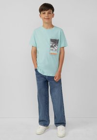 Mintgroene katoenen T-shirt met een grafisch ontwerp in zwart en oranje accenten. Draagt met losse blauwe jeans en witte sneakers.