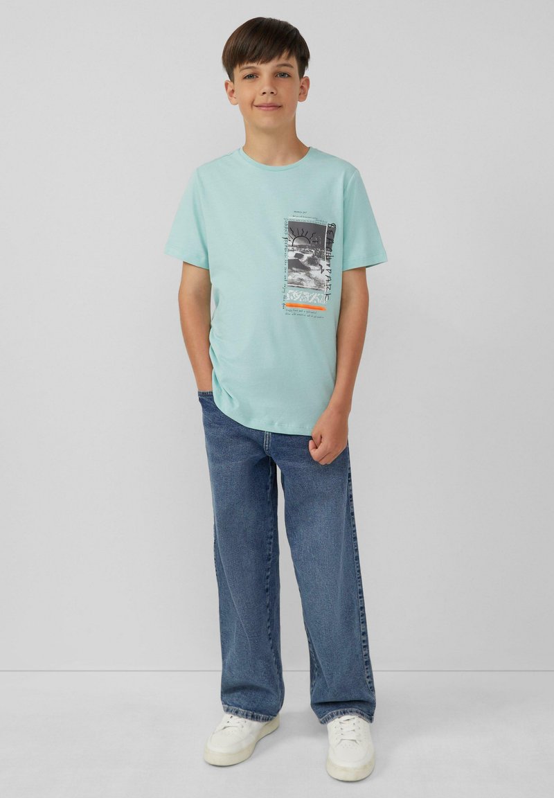 Mintgroene katoenen T-shirt met een grafisch ontwerp in zwart en oranje accenten. Draagt met losse blauwe jeans en witte sneakers.